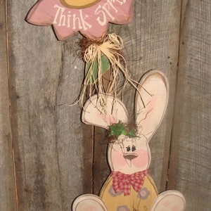 May include: A wooden bunny rabbit with a pink flower on its head. The flower has a yellow center and the words "Think Spring!" written on it. The bunny is wearing a red and white checkered bow tie and has a yellow body with pink flowers.