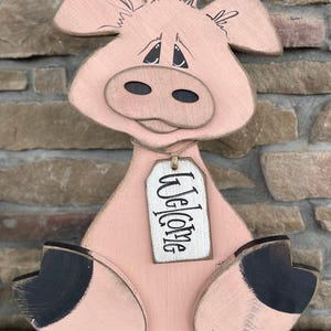 Puede incluir: Una figurita de cerdo de madera rosa con pezuñas negras y una etiqueta blanca que dice "Bienvenido".
