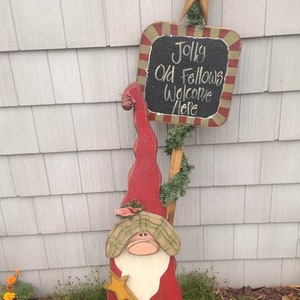 May include: A wooden Christmas gnome decoration with a red hat, white beard, and plaid accents. The gnome is holding a gold star and is standing next to a sign that reads "Jolly Old Fellows Welcome Here".