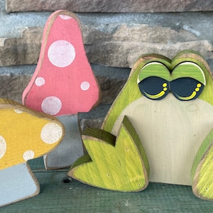 May include: Three wooden figurines: a green frog with large eyes, a yellow mushroom with white polka dots, and a pink mushroom with white polka dots.