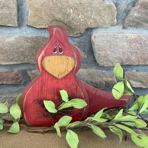 May include: A red wooden cardinal bird figurine with a brown beak and painted eyes. The bird is holding a nest made of twigs and brown twine. The bird is sitting on a brown burlap surface in front of a stone wall.