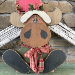Puede incluir: Figura de alce de madera con sombrero rojo, lazo a cuadros rojos y blancos y campana. El alce tiene ojos grandes y oscuros y una bufanda a cuadros rojos y blancos. El alce está sentado sobre una base verde.