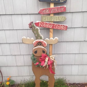 May include: A wooden moose cutout with a red bow and a green wreath around its neck. The moose is holding a sign that says "North Pole" with arrows pointing to "Santa's Workshop", "Elf Village", and "Reindeer Stables".