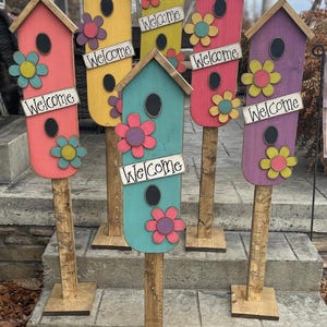 Peut inclure: Six nichoirs à oiseaux en bois peints dans différentes couleurs, dont le rose, le jaune, le bleu, le violet et le vert. Chaque nichoir a une pancarte d'accueil et un motif floral. Les nichoirs sont sur des poteaux en bois et sont disposés en rangée sur un escalier.