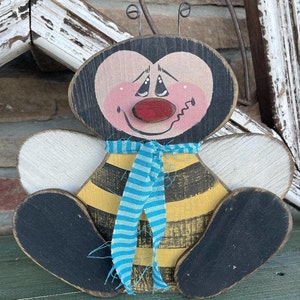 May include: A wooden bee figurine with a black and yellow striped body, white wings, and a painted face. The bee is wearing a blue and white striped ribbon around its neck.