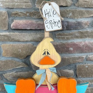May include: A wooden duck holding an umbrella with a pink and blue design. The duck is wearing a blue and orange checkered bow. The umbrella has a sign that says "Hello Spring".