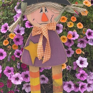 May include: A wooden witch figurine with a black hat, purple dress, orange and yellow striped legs, and a green and brown bow. The figurine is standing in front of a bed of purple, yellow, and orange flowers.