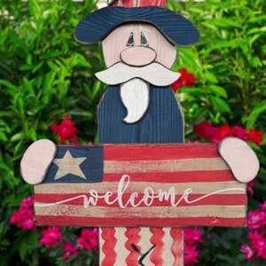 May include: Wooden patriotic figure of a person wearing a blue hat with a white star, a red, white, and blue striped shirt, and red, white, and blue striped pants. The figure is holding a sign that says "welcome" in white lettering.