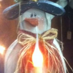 May include: A snowman figurine with a black top hat, a red and white bow, and a lit candle. The snowman is made of wood and has a rustic, vintage look.