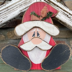 May include: A wooden Santa Claus figurine with a red hat, white beard, and black boots. The figurine is painted with a whimsical style and has a cheerful expression.