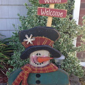 Pode incluir: Decoração de boneco de neve de madeira com uma echarpe xadrez vermelha e preta, um chapéu-coco preto e um nariz de cenoura. O boneco de neve está segurando uma pequena luz. A placa acima diz "Winter Welcome".