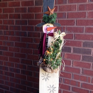 May include: A wooden snowman decoration with a black hat, red scarf, and a small green tree in its arms. The snowman is standing on a white base with snowflakes painted on it.