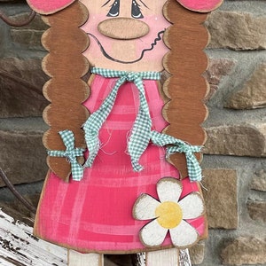 May include: Wooden folk art figure of a girl in a pink dress and hat, with a daisy accent. The figure has brown hair and striped legs. A charming, whimsical decoration.