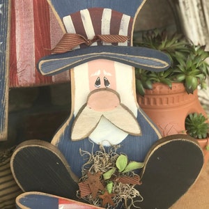 May include: A wooden figurine of a person wearing a blue and white hat with a red, white, and blue ribbon. The figurine is painted in red, white, and blue colors and has a white beard and mustache. The figurine is holding a small bouquet of green plants and is sitting on a brown surface.
