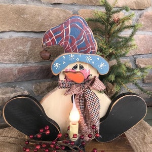 Könnte beinhalten: Eine Holzfigur eines Schneemanns mit einem blauen und roten karierten Hut, einem weißen Schal mit rot-schwarzem kariertem Besatz und schwarz bemalten Füßen. Der Schneemann hält eine brennende Kerze in seinen Händen. Die Figur steht auf einem Holzsockel mit roten Beeren und Grün.