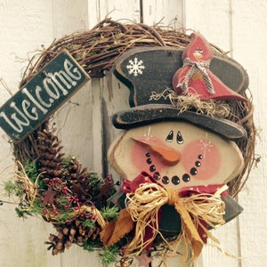 May include: A wooden snowman wreath with a black top hat, red bow, and a welcome sign. The wreath is decorated with pine cones, greenery, and a red cardinal.