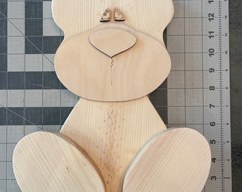 May include: Unfinished wooden bear cutout with a nose and eyes. The bear is sitting with its legs outstretched.