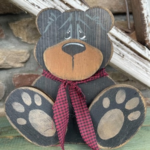 May include: A wooden black bear figurine with a red and black checkered scarf. The bear has a sad expression and is sitting with its paws out.