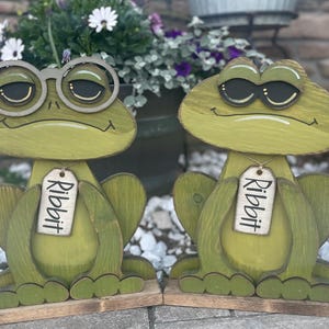 May include: Two wooden frog figurines with a distressed green finish. One frog wears glasses, the other sunglasses. Each frog has a tag that reads "Ribbit". The frogs are on wooden bases, suitable for garden decor.