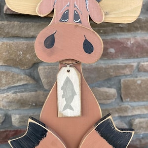May include: A wooden moose figurine with a distressed finish. The moose is painted in shades of brown and tan, with black accents on the hooves. A small tag with a fish silhouette hangs from its neck. The moose has a whimsical, folk art style.