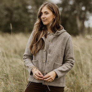 May include: A woman wearing a brown hooded jacket with a button closure, standing in a field of tall grass. She is looking off to the side with a thoughtful expression.