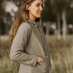 May include: A woman wearing a gray hooded jacket with a button closure and a patch pocket. She is standing in a field of tall grass.