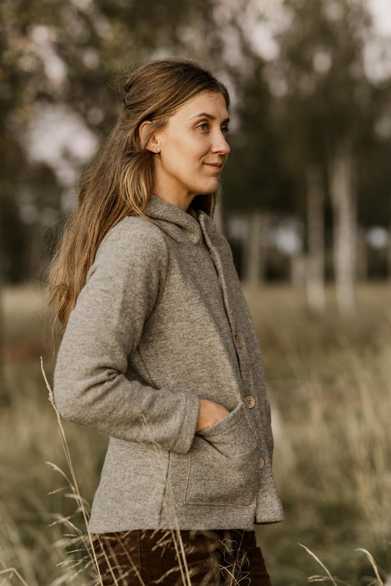 May include: A woman wearing a gray hooded jacket with a button closure and a patch pocket. She is standing in a field of tall grass.