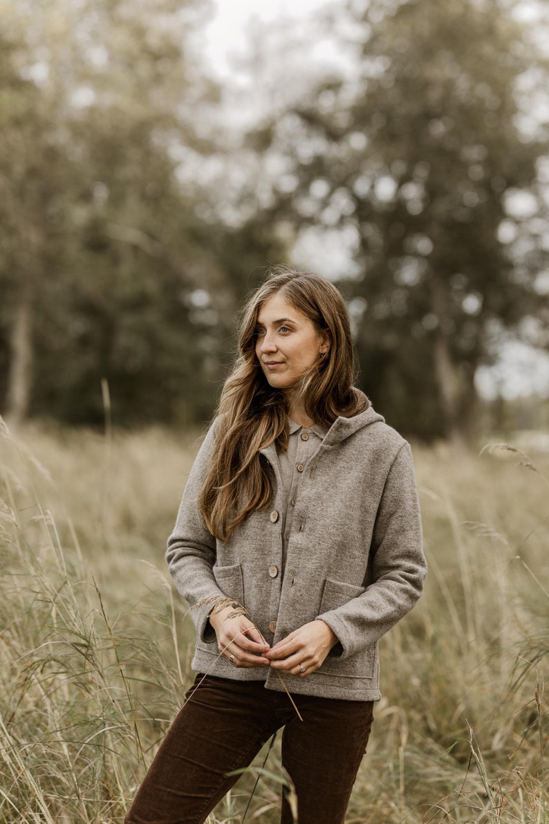 May include: A woman wearing a brown hooded jacket with a button closure, standing in a field of tall grass. She is looking off to the side with a thoughtful expression.