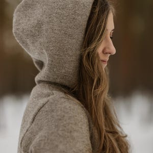 May include: A woman wearing a gray hooded sweater with her hair down. She is looking down and her face is partially obscured by the hood.