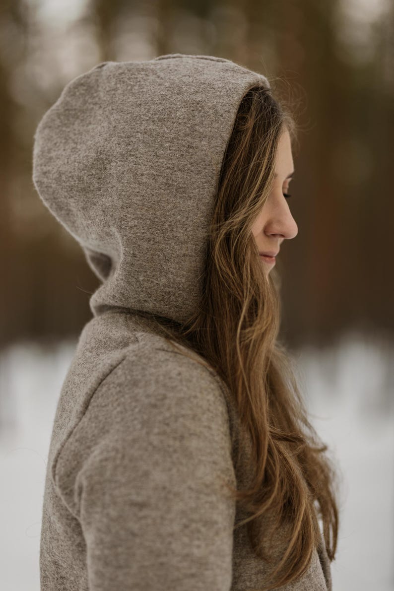 May include: A woman wearing a gray hooded sweater with her hair down. She is looking down and her face is partially obscured by the hood.