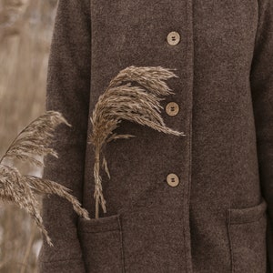 May include: A brown wool coat with a stand-up collar and wooden buttons. The coat has two patch pockets and is worn by a person standing in a field of tall grass.