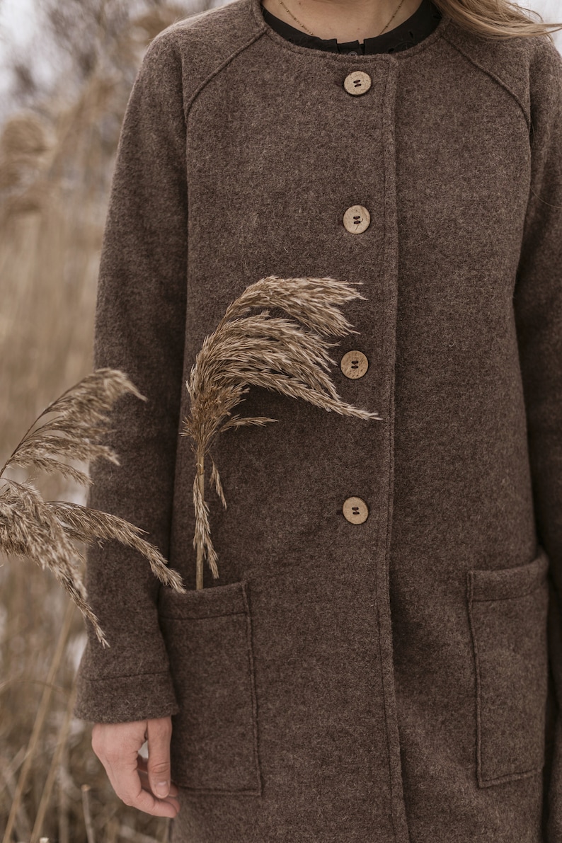May include: A brown wool coat with a stand-up collar and wooden buttons. The coat has two patch pockets and is worn by a person standing in a field of tall grass.