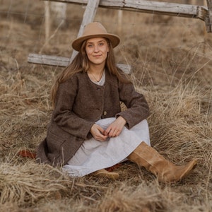 May include: A woman wearing a brown coat and a white dress sits in a field of dry grass. She is wearing a brown hat and brown boots.