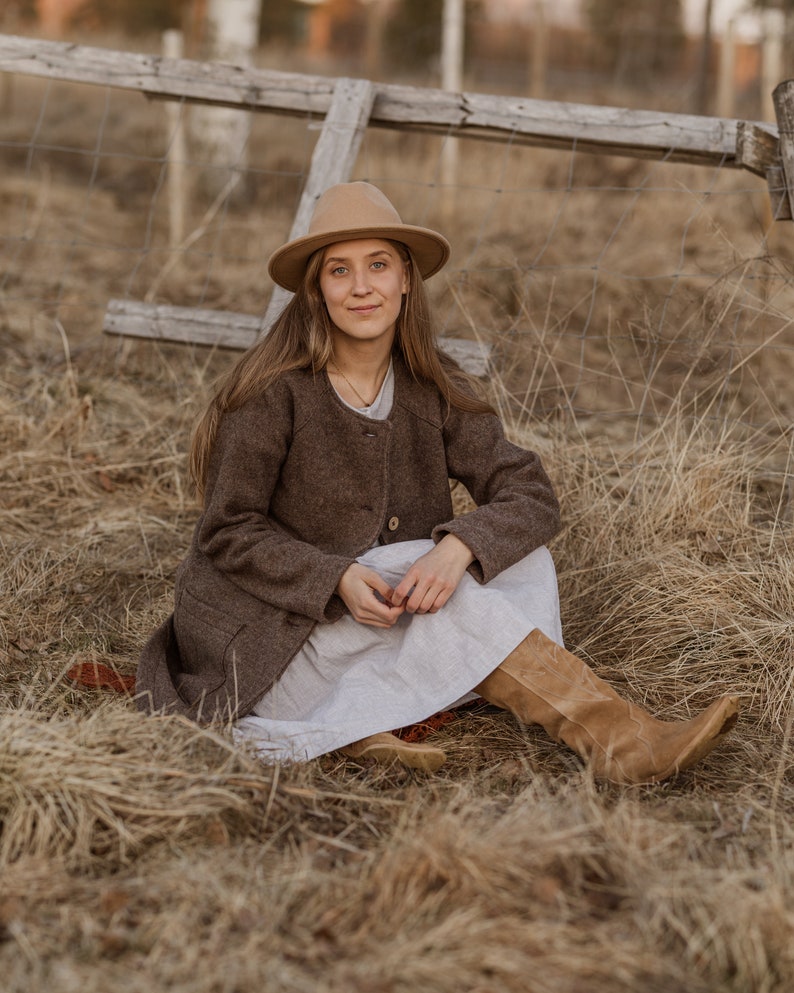May include: A woman wearing a brown coat and a white dress sits in a field of dry grass. She is wearing a brown hat and brown boots.