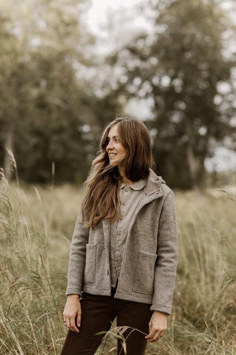 May include: A woman wearing a gray hooded jacket with a brown button-up shirt underneath. She is standing in a field of tall grass.