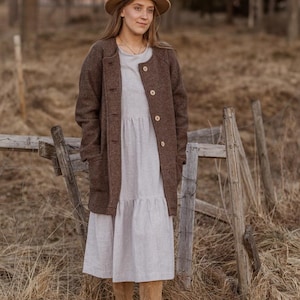 May include: A woman wearing a brown wool coat with a white dress and brown boots. She is standing in a field with a wooden fence behind her.