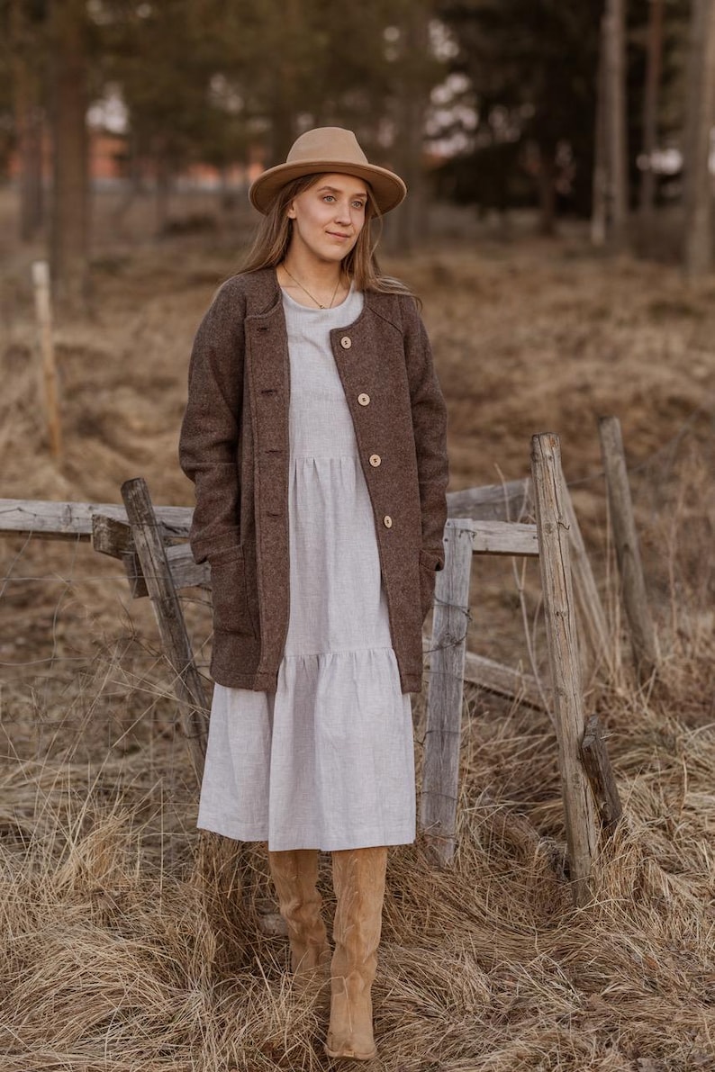 May include: A woman wearing a brown wool coat with a white dress and brown boots. She is standing in a field with a wooden fence behind her.