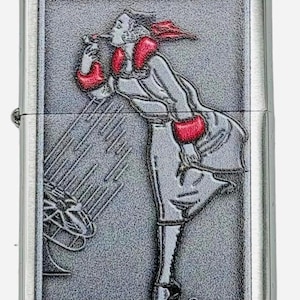 May include: A silver-colored lighter with a detailed embossed design. The design features a woman in a long coat and red accents, blowing into a stylized object. The lighter has a rectangular shape with a hinged lid.