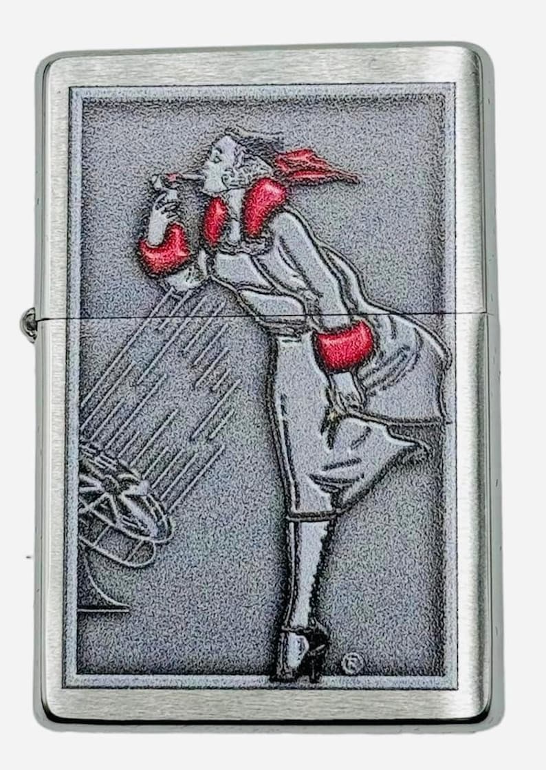 May include: A silver-colored lighter with a detailed embossed design. The design features a woman in a long coat and red accents, blowing into a stylized object. The lighter has a rectangular shape with a hinged lid.