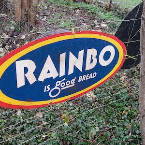 May include: A vintage oval-shaped metal sign with red, yellow, and blue paint. The sign advertises "Rainbo is good bread".