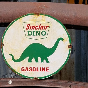 Puede incluir: Un letrero redondo de metal blanco con una silueta de dinosaurio verde y el texto "Sinclair Dino Gasoline" en letras rojas.