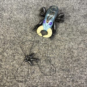 May include: A plastic beetle toy featuring a metallic blue and black body with purple eyes and large, cream-colored mandibles. The toy has six black legs and is connected by a thin black string to a small, black, three-pronged plastic accessory.