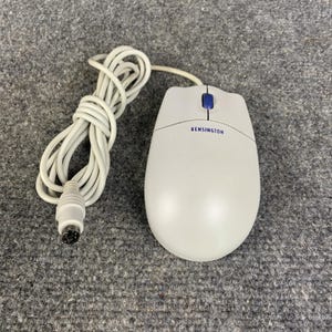 May include: A light gray Kensington computer mouse with a blue scroll wheel and a long, coiled light gray cord ending in a round PS/2 connector. The brand name Kensington is printed in blue text on the top surface of the mouse body.