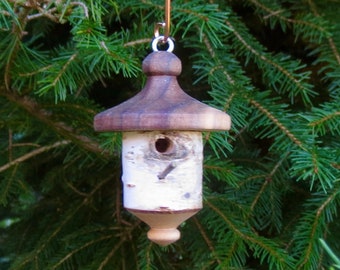 Könnte beinhalten: Ein kleiner Vogelhaus-Ornament aus Holz mit braunem Dach und weißem Birkenrindenkörper. Das Vogelhaus hängt an einem dünnen, kupferfarbenen Draht vor einem Hintergrund aus grünen Kiefernnadeln.