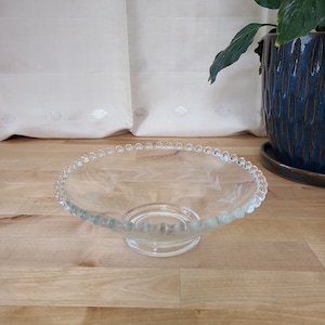 Puede incluir: Un cuenco de vidrio transparente con un borde de cuentas y un diseño floral grabado. El cuenco tiene una base de pedestal.