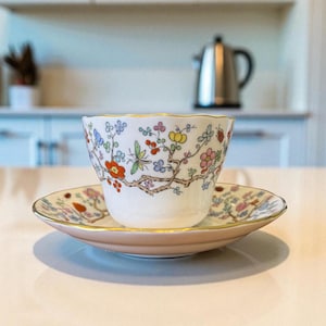 May include: A white teacup and saucer set with a floral design. The cup and saucer have a pink and gold trim. The floral pattern includes red, yellow, and pink flowers with green leaves. The set is on a white surface.