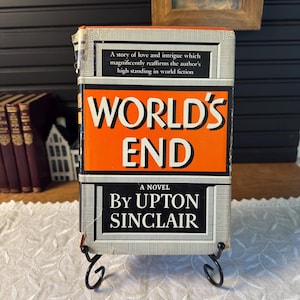 May include: A vintage book titled "World's End" by Upton Sinclair. The book has an orange cover with white and black text. The title is prominently displayed. The book is propped up on a black metal stand.