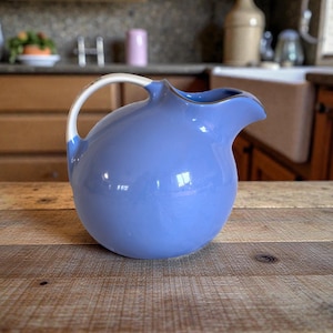 May include: A light blue ceramic pitcher with a white handle and a gold rim. The pitcher is sitting on a wooden table. The background is a kitchen with wooden cabinets and a sink.