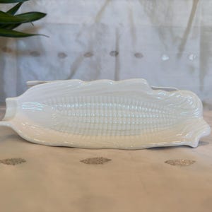 May include: White ceramic corn on the cob serving dish. The dish is shaped like an ear of corn and has a textured surface.