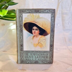 May include: Antique book titled "The Temptation of Olive Latimer" by Mrs. L.T. Meade. The cover features a portrait of a woman wearing a large hat with a feather. The book has a light blue cover with floral and scroll designs.